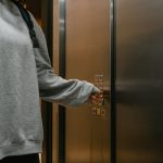 Person in grey sweater pushing elevator button with backpack in a modern building.